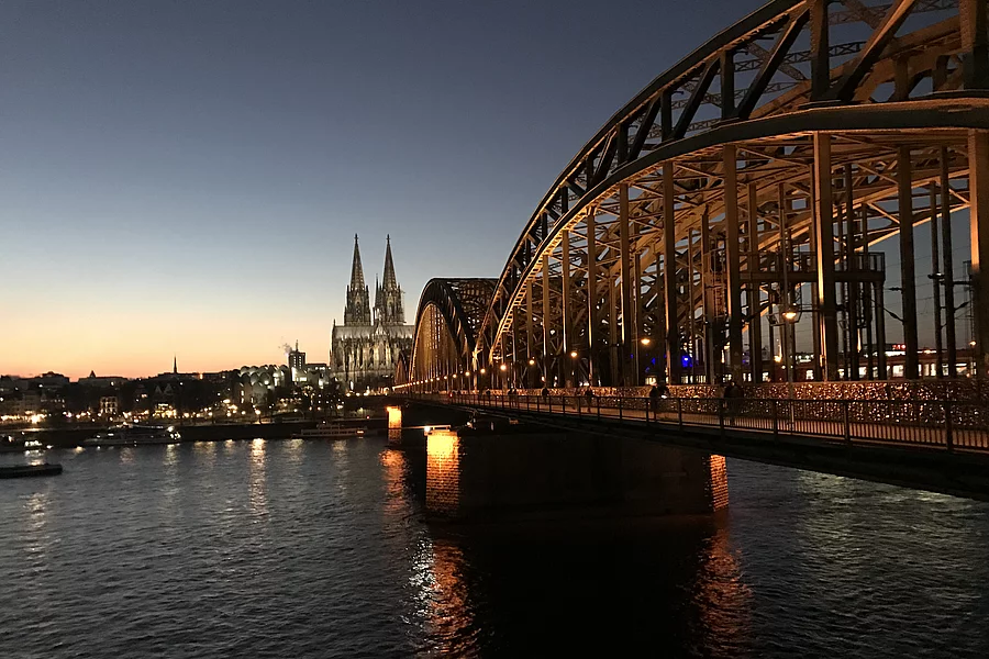 The Hohenzollern Bridge in Cologne is illuminated at night and spans the Rhine. The Cologne Cathedral can be seen in the background.