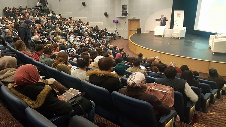 The photo shows a conference hall with participants looking towards a stage on which a speaker is standing, showing a presentation on a screen.